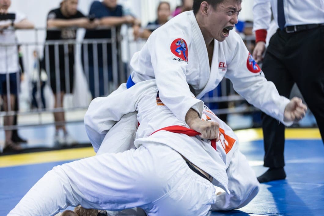 A Brazilian Jiu-Jitsu competitor in a white gi and blue belt pins his opponent to the mat and roars, fist clenched, as the referee looks on.