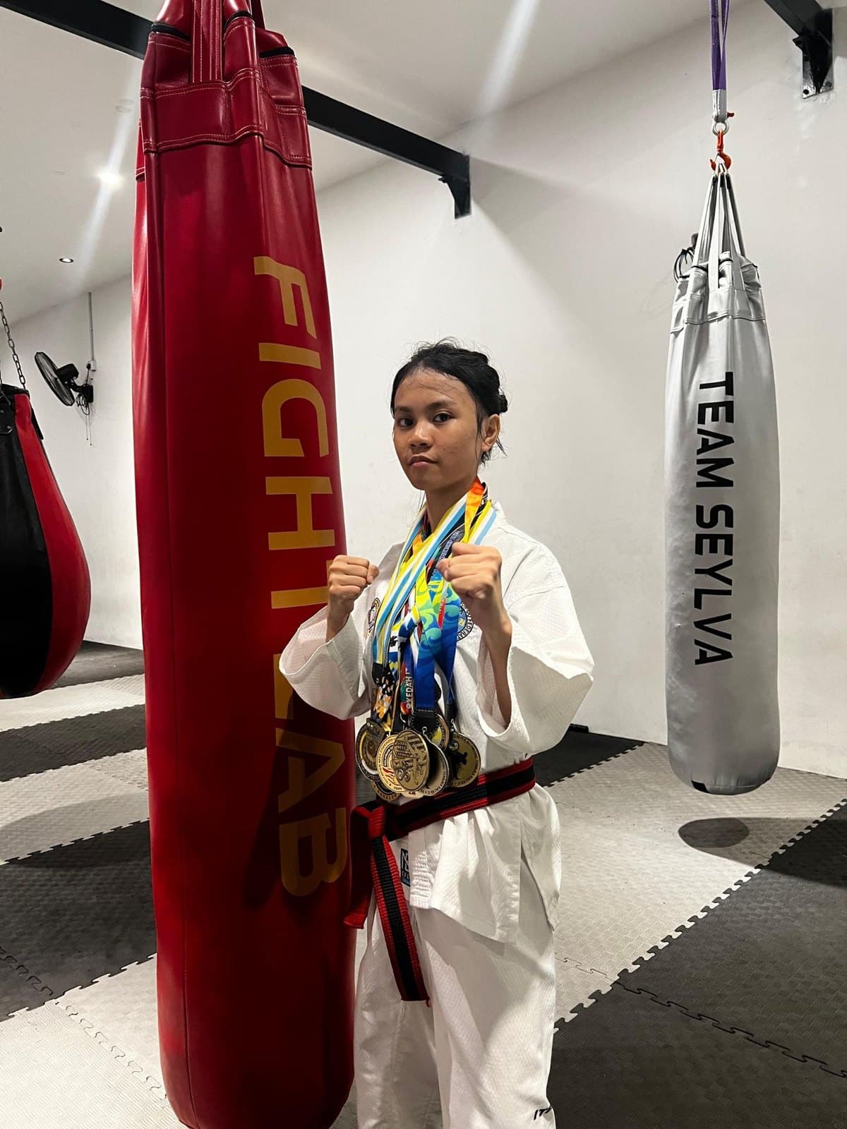 Fightlab student Nur Qaseh Diyani in her Taekwon-Do uniform and black-and-red belt, fists raised, wearing her gold medals beside the Fightlab and Team Seylva punching bags.
