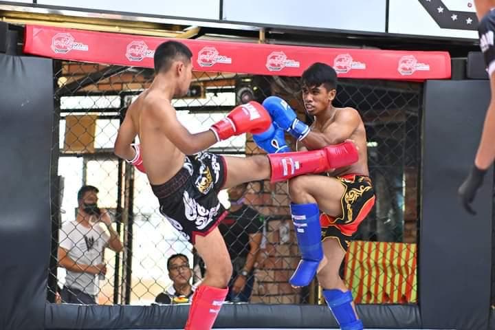 A Muay Thai fighter in black shorts lands a body kick during a cage bout.
