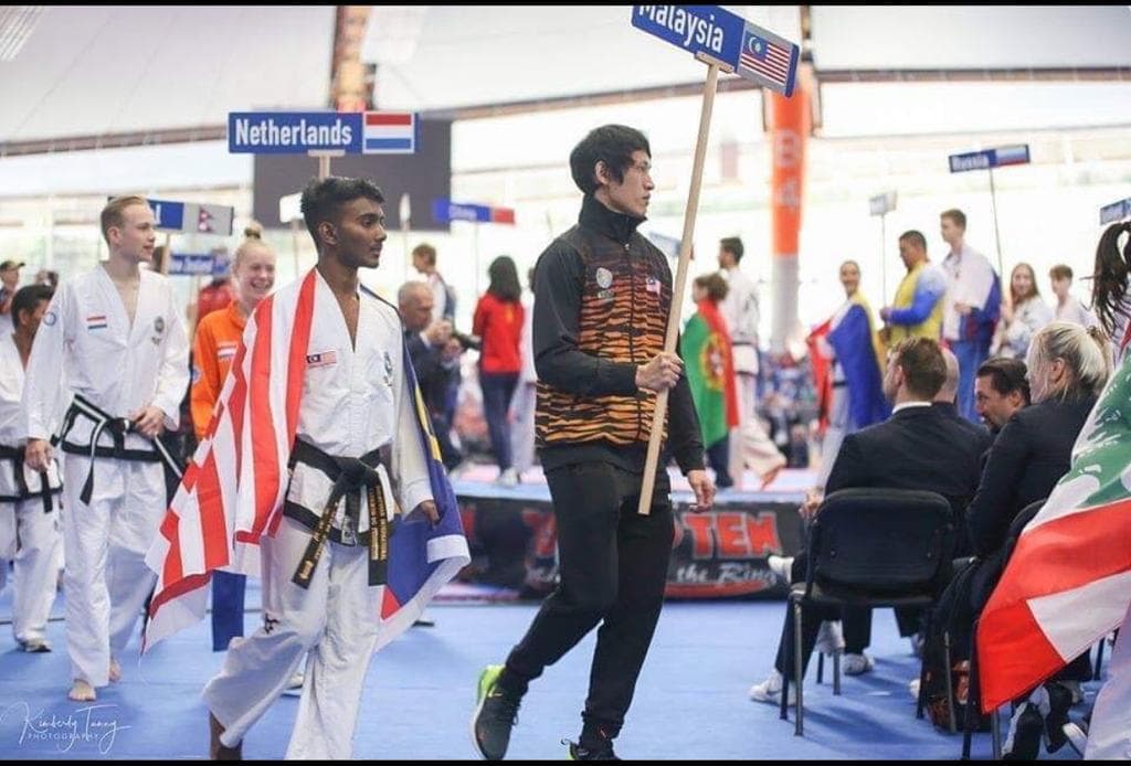A black belt draped in the Malaysian flag walks behind the Malaysia sign in the parade of nations at an international Taekwon-Do event.
