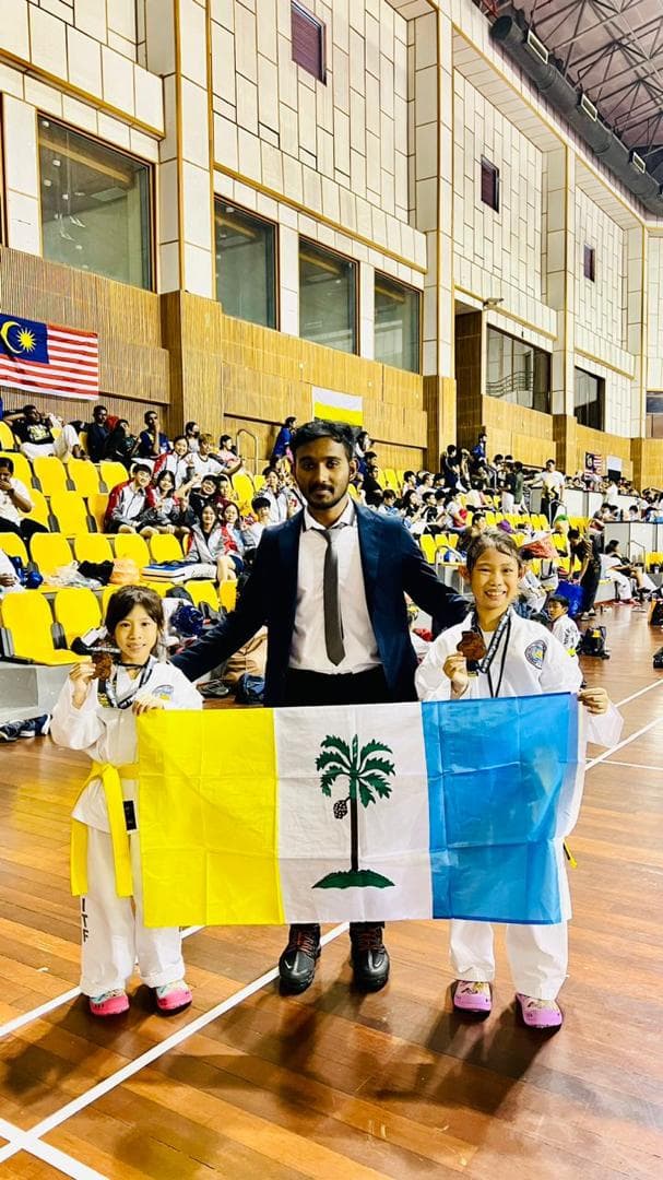 A coach stands with two young Taekwon-Do students holding their medals and the Penang flag in a sports hall.